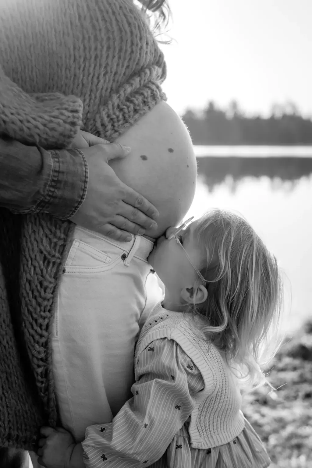 Babybauchshooting Weilheim, Schwangere im Profil auf einer Wiese, das Geschwisterkind küsst den Babybauch
